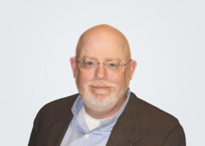 Headshot of David Stout, a man with a white beard, in front of a light grey background.