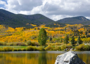 The mountainsides in Summit County, Colorado shimmer with the orange, gold and green colors of autumn. Pictured here are the pine and aspen forests cascading down the mountains, with a lake in the foreground.