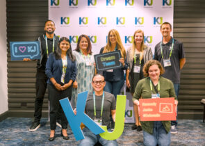 8 KJ employees pose for a picture at a company conference. The employees are men and women of various ages, wearing business casual attire. They stand in front of backdrop with a blue and green KJ logo pattern on it. This image relates to employee ownership.