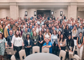 A large group of KJ employees pose for a photo at TechEx 2024. The employees are dressed in business casual clothing and stand in a cream-colored banquet room.