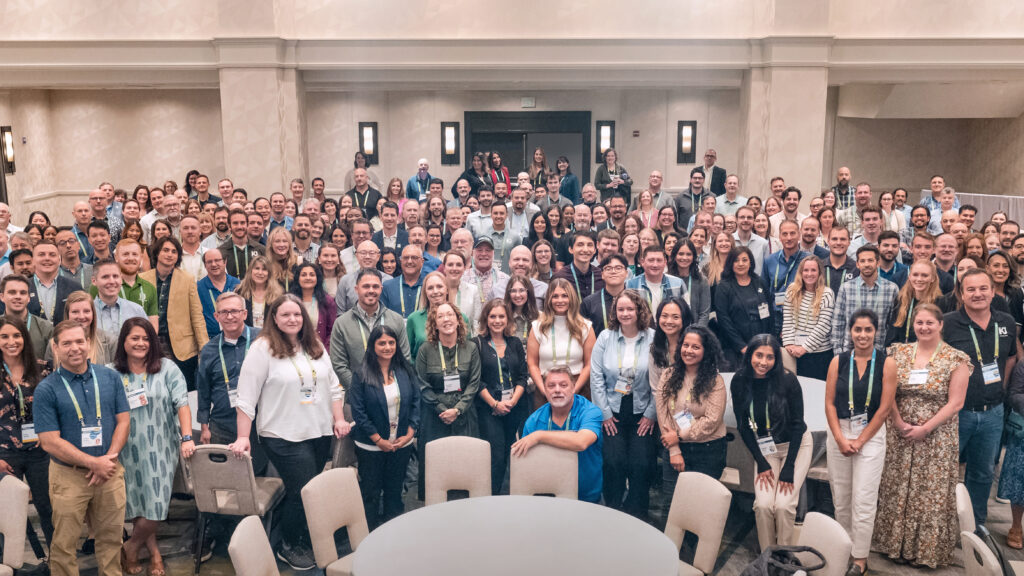 A large group of KJ employees pose for a photo at TechEx 2024. The employees are dressed in business casual clothing and stand in a cream-colored banquet room.